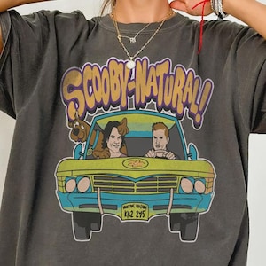 May include: Dark gray t-shirt featuring a cartoon image of the Mystery Machine van with the text "Scooby Natural!". The van has a yellow body, blue accents, and the license plate reads "KAZ 295". The shirt has short sleeves and a relaxed fit.