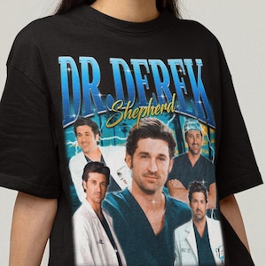 Peut inclure: T-shirt noir avec un collage d'images du Dr. Derek Shepherd et le texte "DR. DEREK Shepherd" en bleu et jaune. Le t-shirt a un col rond et des manches courtes.