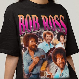 Op de afbeelding: Zwart T-shirt met een retro afbeelding van Bob Ross en de tekst "BOB ROSS" in een roze en oranje verloop. De afbeelding bevat meerdere afbeeldingen van Bob Ross. Het shirt heeft een ronde hals en korte mouwen.