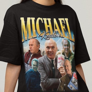 Puede incluir: Camiseta negra con un diseño gráfico de estilo vintage de Michael Keaton y un collage de imágenes. El nombre "MICHAEL Keaton" es dorado con un fondo degradado azul. La camiseta la lleva una persona.