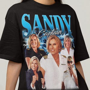 May include: Black t-shirt featuring a graphic of Captain Sandy with the text "SANDY Captain" in blue. The graphic includes multiple images of Captain Sandy. The shirt is worn by a person.