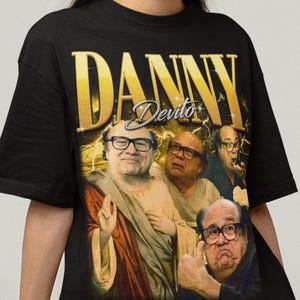 May include: Black t-shirt featuring a graphic of Danny DeVito with the text "DANNY DeVito" in gold lettering. The graphic includes multiple images of DeVito. The shirt is worn by a person.