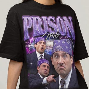 以下が含まれることがあります： 「The Office」のキャラクター「Prison Mike」のグラフィックが特徴の黒いTシャツ。デザインには、紫色の稲妻のアクセントと、紫色のバンダナを身に着けたキャラクターの複数の画像とともに、「PRISON Mike」というテキストが含まれています。