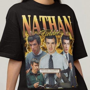 Pode incluir: Camiseta preta com um gráfico de Nathan Fielder em várias poses, com o nome "NATHAN FIELDER" em letras douradas. O design tem uma estética vintage e retrô.