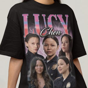May include: Black t-shirt featuring a graphic of the name "Lucy Chen" in pink and blue, with images of a woman. The shirt has a vintage style design.