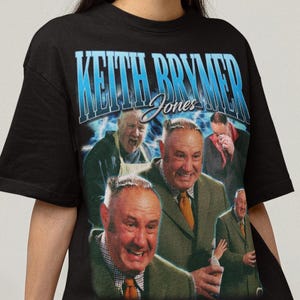 Puede incluir: Camiseta negra con un gráfico retro de un hombre con traje, con el texto "Keith Brymer Jones" en azul y blanco. El gráfico incluye múltiples imágenes del hombre. La modelo lleva pantalones cortos grises.