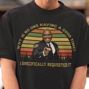 May include: Black t-shirt with a graphic featuring a man pointing, set against a sunset design with the text "WHY IS NO ONE HAVING A GOOD TIME? I SPECIFICALLY REQUESTED IT."