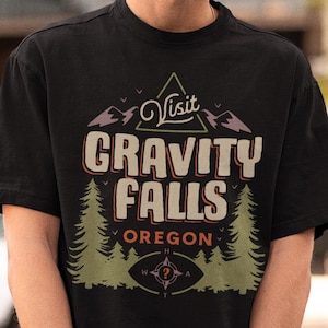 Könnte beinhalten: Schwarzes T-Shirt mit einem Grafikdesign mit der Aufschrift "Visit Gravity Falls Oregon". Das Design umfasst Berge, Bäume und einen Kompass. Das T-Shirt hat kurze Ärmel.