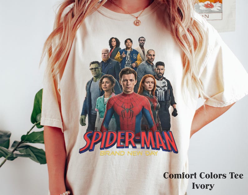 Op de afbeelding: Ivoor Comfort Colors T-shirt met een afbeelding van Spider-Man en andere personages. De tekst "SPIDER-MAN BRAND NEW DAY" is in rood en blauw. Het shirt heeft een ronde hals.