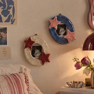 May include: Two oval-shaped ceramic wall frames with glossy finishes. One frame is speckled cream with two dark red star accents, and the other is mottled blue with two light pink star accents. Each frame holds a small black and white photograph.