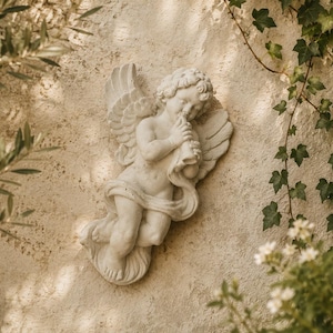 Puede incluir: Una escultura de querubín blanco tocando una trompa, montada en una pared beige texturizada. El querubín tiene alas detalladas y está rodeado de vegetación, incluyendo ramas de olivo e hiedra, creando una estética serena y clásica.