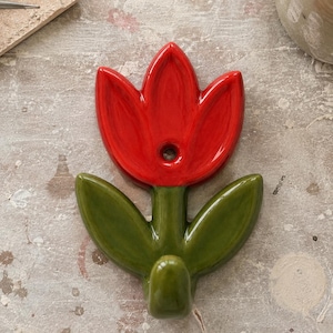 Puede incluir: Un gancho de pared de cerámica brillante con forma de flor de tulipán roja con dos hojas verdes que se extienden desde el tallo. El centro del pétalo del tulipán rojo presenta un orificio circular para el montaje y la base del tallo se curva.