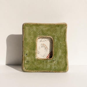 May include: A square ceramic picture frame with a glossy, mottled olive green glaze and an uneven, hand-formed edge. The frame features a central rectangular opening containing a small black and white line drawing of a person's profile.