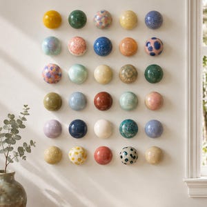 Może przedstawiać: Kolekcja trzydziestu okrągłych ceramicznych dekoracji ściennych ułożonych w siatkę. Każdy element posiada unikalne szkliwo, w tym jednolite kolory, takie jak żółty, zielony, niebieski i czerwony, a także nakrapiane, kropkowane i marmurkowe wzory.