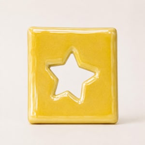 Pode incluir: Um objeto quadrado de cerâmica com acabamento amarelo-mostarda brilhante, apresentando um recorte em forma de estrela no centro. A peça possui bordas arredondadas e uma superfície lisa e reflexiva, com cor uniforme em toda a face frontal.