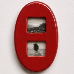 Könnte beinhalten: Ein glänzender, ovaler Bilderrahmen aus Keramik in Rot mit zwei rechteckigen Öffnungen. Die obere Öffnung zeigt ein schwarz-weißes Landschaftsfoto einer Küste, während die untere Öffnung ein schwarz-weißes Porträt einer Person zeigt.