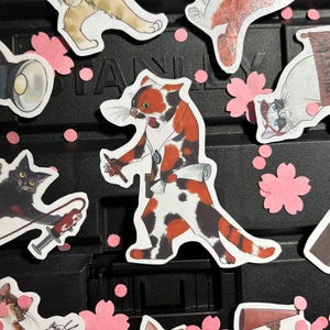 Puede incluir: Una colección de pegatinas de gatos con varios diseños, incluyendo un gato calicó sosteniendo un pergamino y una jeringa. Las pegatinas presentan una mezcla de colores, incluyendo naranja, negro y blanco, con acentos de flores de cerezo rosas. Las pegatinas están sobre una superficie negra.