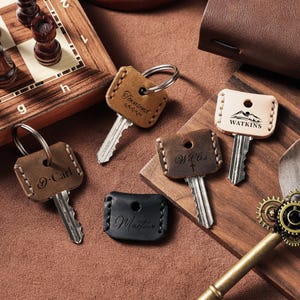 Puede incluir: Una colección de llaveros de cuero personalizados en varios tonos de marrón y negro. Cada llavero tiene una llave y está grabado con un nombre o palabra. Los llaveros están dispuestos sobre una superficie de madera, con un juego de ajedrez al fondo.