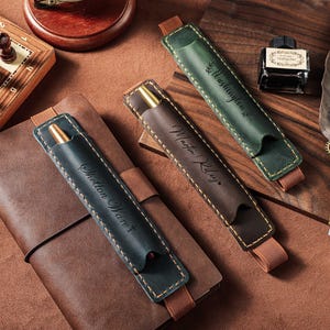 May include: An overhead shot showcases a chess set, a leather-bound journal, and several leather pen holders in shades of green and brown. The pen holders are embossed with names. A decorative quill pen and ink bottle complete the scene.