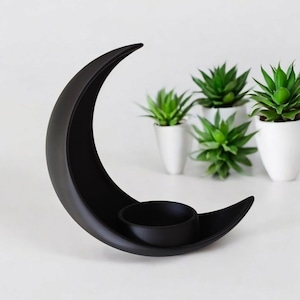May include: A black, crescent moon-shaped candle holder with a circular cup for a candle. The holder is made of a matte black material. Small potted green succulents are in the background, creating a minimalist aesthetic.