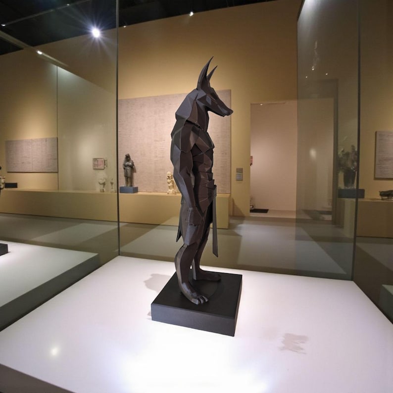 May include: A dark gray, geometric sculpture of a standing jackal-headed figure, reminiscent of Anubis, featuring sharp, angular facets and a smooth matte finish. The figure is mounted on a rectangular black base and displayed in a museum setting.