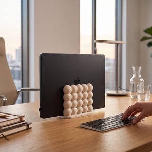 Op de afbeelding: Een matzwarte laptop wordt rechtop gehouden door een verticale standaard met een raster van crèmekleurige bollen. De standaard heeft een rechthoekige voet en ondersteunt het apparaat in een gesloten positie voor op het bureau.