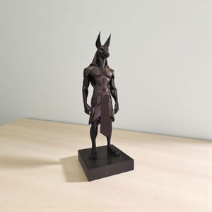 May include: A dark brown, low-poly geometric sculpture of the Egyptian deity Anubis standing in a rigid, upright posture. The figure features sharp, angular facets, pointed ears, and a simple loincloth, all mounted on a solid rectangular base.