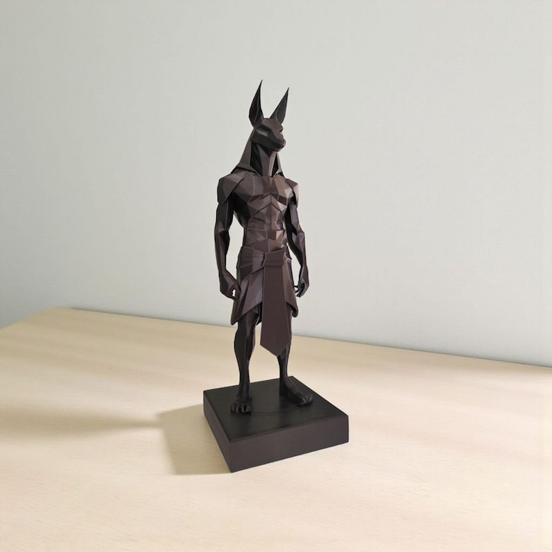 May include: A dark brown, low-poly geometric sculpture of the Egyptian deity Anubis standing in a rigid, upright posture. The figure features sharp, angular facets, pointed ears, and a simple loincloth, all mounted on a solid rectangular base.