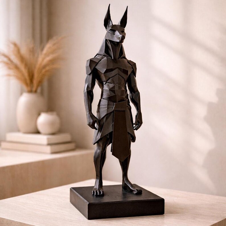 May include: A matte black, geometric-style sculpture of the Egyptian deity Anubis, depicted with a jackal head and human-like muscular torso. The figure stands on a rectangular black base, featuring sharp, angular facets throughout its form.