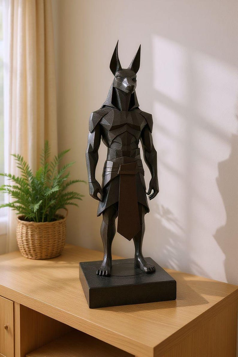 May include: A dark charcoal gray, geometric-style statue of the Egyptian deity Anubis, featuring a jackal head, muscular human-like torso, and a traditional kilt. The figure stands upright on a solid, rectangular black base with a matte finish.