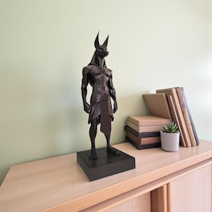 May include: A dark brown, low-poly geometric sculpture of the Egyptian deity Anubis, featuring a jackal head and human-like physique. The figure stands on a rectangular base, showcasing sharp, angular facets and a smooth, matte finish throughout.