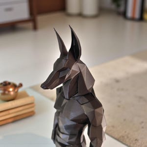 May include: A dark brown, low-poly geometric sculpture of the Egyptian deity Anubis, featuring a stylized canine head with tall, pointed ears and a muscular torso. The figure is constructed from multiple flat, angular planes.