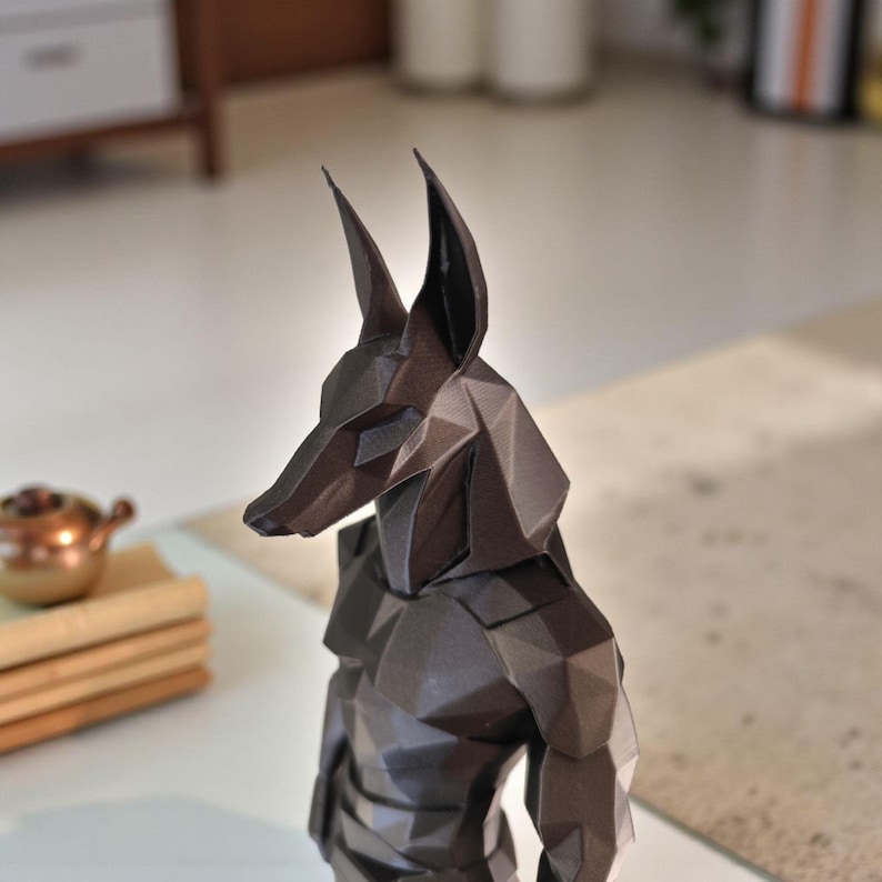 May include: A dark brown, low-poly geometric sculpture of the Egyptian deity Anubis, featuring a stylized canine head with tall, pointed ears and a muscular torso. The figure is constructed from multiple flat, angular planes.