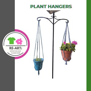 Puede incluir: Dos macetas colgantes suspendidas de un soporte de metal negro. Una maceta es azul, la otra es de terracota, ambas llenas de vegetación y flores. El texto "PLANT HANGERS" está en la parte superior. El logotipo "RE-ART: UPCYCLED FASHION" está a la izquierda.