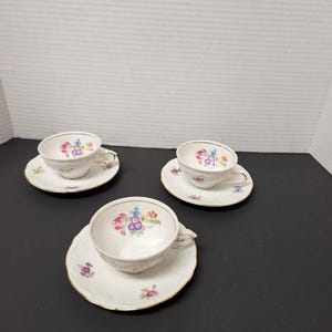 May include: Three white teacups and saucers with floral designs. Each cup and saucer has a gold trim. The floral pattern includes pink, purple, blue, and yellow flowers. The teacups have small handles.
