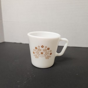 May include: White ceramic mug with a brown floral design. The mug has a small handle and a slightly flared rim. The floral pattern includes small flowers and leaves. The mug is likely vintage and suitable for coffee or tea.