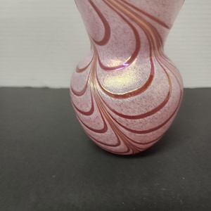 Op de afbeelding: Een kleine, roze glazen vaas met een bolle basis en een uitlopende bovenkant. De vaas heeft een wervelend patroon van roodbruine lijnen tegen een gespikkelde achtergrond. Het glas heeft een iriserende glans, wat een subtiele glans toevoegt.