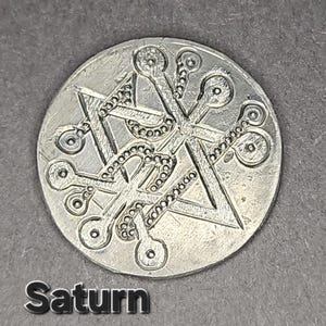 May include: A circular silver-toned metal coin featuring an engraved geometric design of intersecting lines, triangles, and circular terminals. The word Saturn is printed in a bold, sans-serif font at the bottom left of the image.