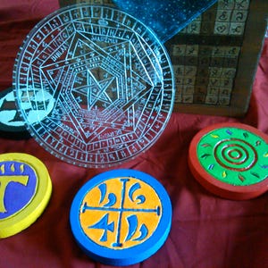 Puede incluir: Una colección de objetos circulares decorativos con símbolos místicos. Incluye un círculo de vidrio transparente grabado con un diseño de estrella y varios posavasos de madera pintados en varios colores y patrones. Una caja de madera con símbolos tallados está en el fondo.