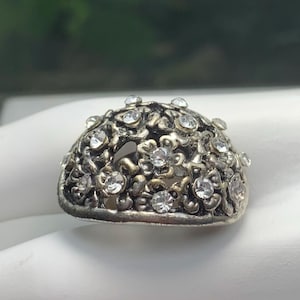 May include: A silver-toned ring with a floral design, encrusted with clear, round gemstones. The ring has an openwork design, with the gemstones set within the floral pattern. The ring is on a white surface.
