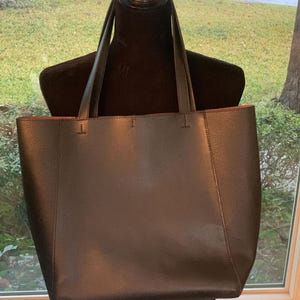 May include: A black tote bag with long handles, displayed on a black mannequin. The bag has a textured appearance and a contrasting red interior. The bag is rectangular and appears to be made of leather or a leather-like material.