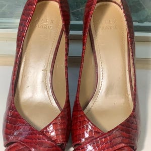 May include: Pair of red, open-toe pumps with a textured, reptile-like pattern. The heels are mid-height, and the shoes have a crisscross design over the foot. The brand name "ALEX MARIE" is visible inside the shoe.