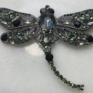 May include: A dragonfly brooch with a silver-tone metal frame. The wings are adorned with clear, black, and green crystals. The body features a large, oval-shaped crystal. The dragonfly's tail is a line of green crystals.