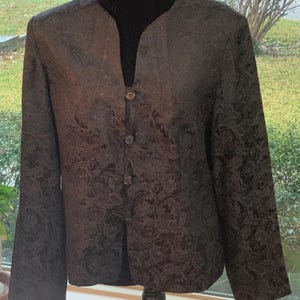 May include: A dark brown blazer with a paisley pattern. The blazer has a notched collar and three buttons down the front. The sleeves are long and the fabric has a subtle sheen. The blazer is displayed on a black mannequin.