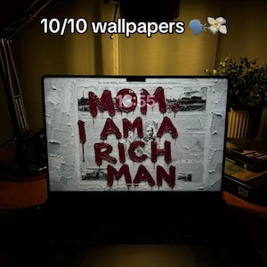 May include: A laptop screen displaying a digital wallpaper with the text 10/10 wallpapers and a money bag emoji. The screen shows a black and white newspaper background with the red, textured, dripping text MOM, I AM A RICH MAN.