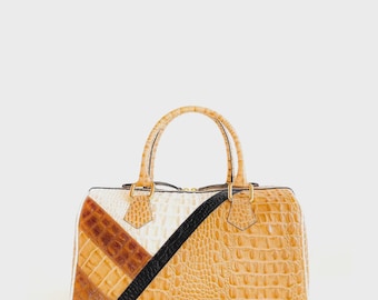 May include: A tan, white, and brown leather handbag with a geometric design. The bag features a black diagonal stripe and a textured, crocodile-like pattern. It has two handles and gold-colored hardware.