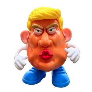 Könnte beinhalten: Ein neuartiges Spielzeug, das einer politischen Figur ähnelt, mit einem orangefarbenen Gesicht, gelbem Haar und blauen Schuhen. Das Spielzeug hat übertriebene Gesichtszüge, darunter große Lippen und einen strengen Ausdruck. Weiße Handschuhe und Arme vervollständigen das Design.