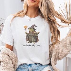 K&ouml;nnte beinhalten: Wei&szlig;es T-Shirt mit einer Illustration eines Frosch-Zauberers, der einen Stab und ein Buch h&auml;lt. Der Frosch tr&auml;gt einen braunen Zaubererhut und Umhang. Der Text "'Twas The Brain Fog M'Lord" ist darunter gedruckt.