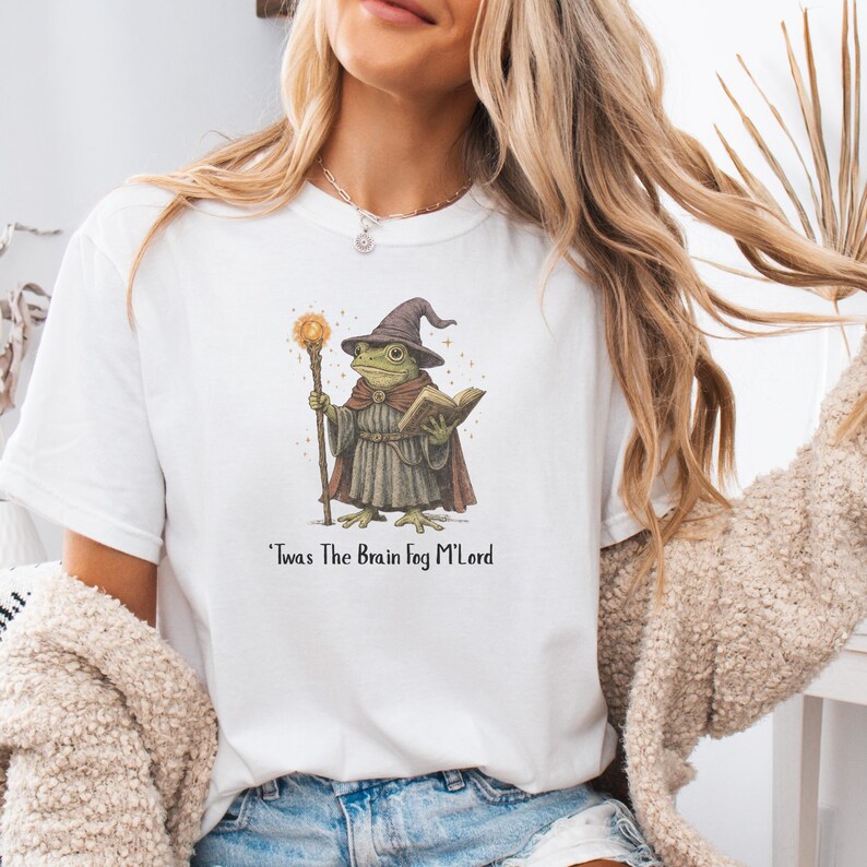 K&ouml;nnte beinhalten: Wei&szlig;es T-Shirt mit einer Illustration eines Frosch-Zauberers, der einen Stab und ein Buch h&auml;lt. Der Frosch tr&auml;gt einen braunen Zaubererhut und Umhang. Der Text "'Twas The Brain Fog M'Lord" ist darunter gedruckt.