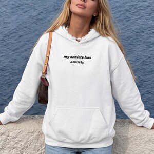 Peut inclure: Un sweat-shirt &agrave; capuche blanc &agrave; manches longues avec le texte noir "my anxiety has anxiety" imprim&eacute; sur la poitrine. Le v&ecirc;tement comprend une poche kangourou &agrave; l'avant, une capuche &agrave; cordon et des poignets c&ocirc;tel&eacute;s, avec un sac &agrave; bandouli&egrave;re marron.